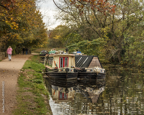 Two boats side by side moored on the Leeds and Liverpool Canal between Hirst Lock and Saltaire in early autumn with a young woman in a pink coat walking into the distance on the towpath