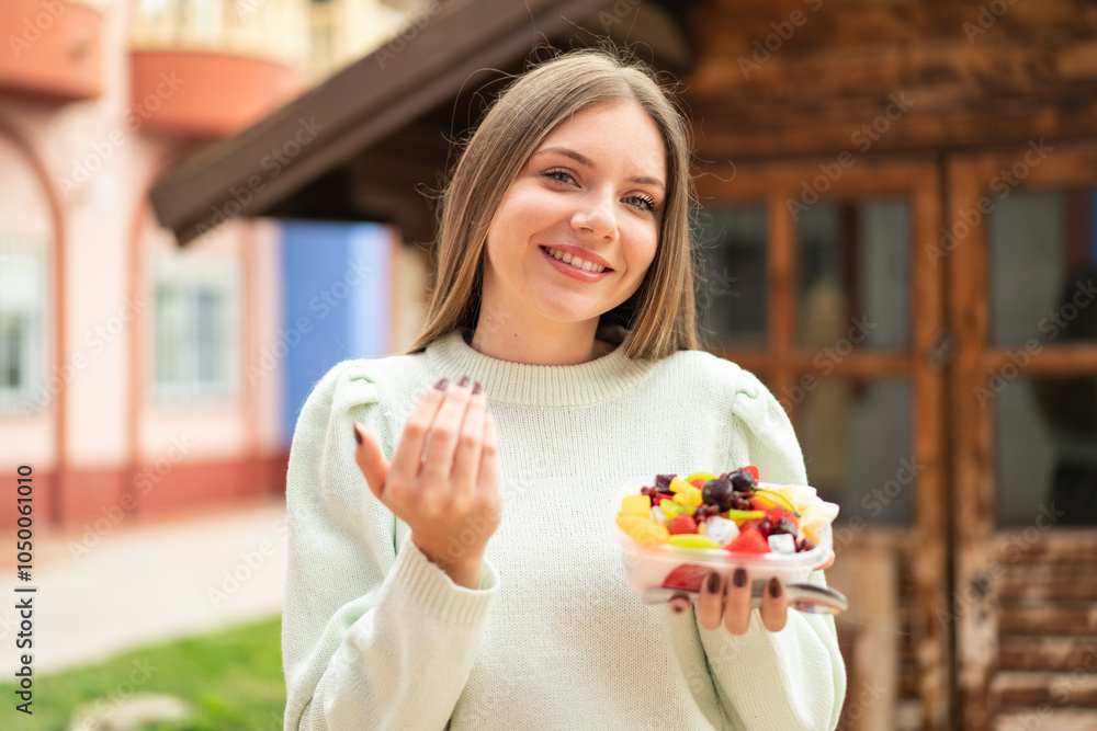 Young pretty blonde woman holding a bowl of fruit at outdoors inviting to come with hand. Happy that you came Young pretty blonde woman holding a bowl of fruit at outdoors inviting to come with hand. Happy that you came