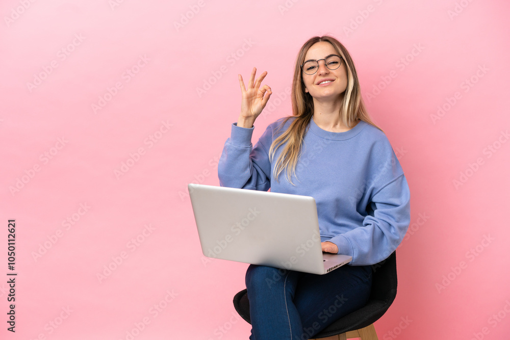Young woman sitting on a chair with laptop over isolated pink background showing ok sign with fingers Young woman sitting on a chair with laptop over isolated pink background showing ok sign with fingers