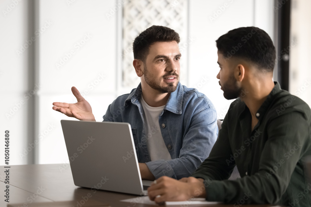 Fototapeta premium Confident young Middle Eastern startup leader man talking to business partner at laptop, explaining marketing task for project, speaking, meeting with colleagues for teamwork at office table