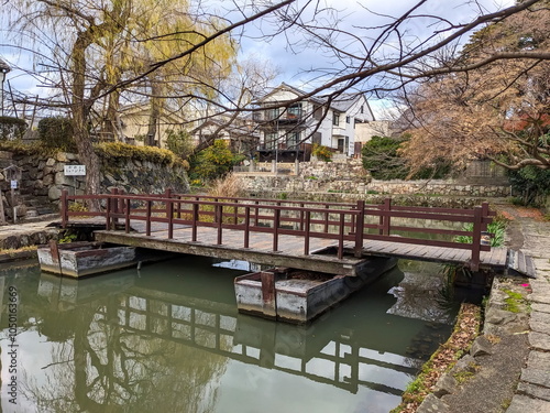 Omihachiman, Japan - 12.20.2023: A simple boat built with two moored boats over Hachimanbori canal alongside footpaths, historic buildings, and autumn trees under a cloudy blue sky without tourists photo