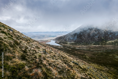 Rolling hills covered in grass lead down to a meandering river reflecting the cloudy sky, creating a peaceful and beautiful natural setting in a valley. photo