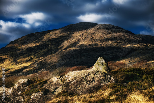 A large, gray rock sits on a hill covered in short brown grass and dry vegetation. The sky is overcast with dark clouds. photo