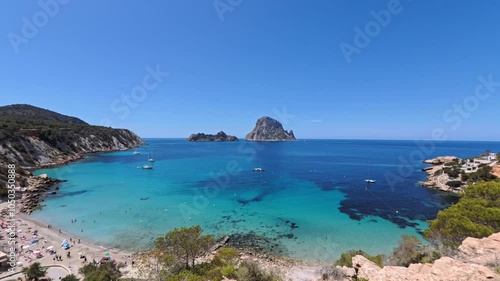 Stunning panorama of Ibiza Cala Hort beach, with Es Vedra and Es Vedranell islands visible in the distance Yachts float on the clear waters, while beachgoers relax on the sandy shore