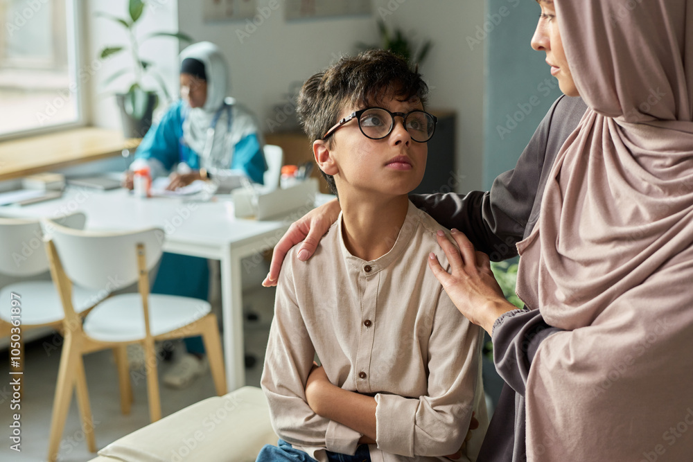 Obraz premium Muslim woman gently reassuring her son in warm, inviting living room as another family member stays in background at a dining table, creating a peaceful atmosphere