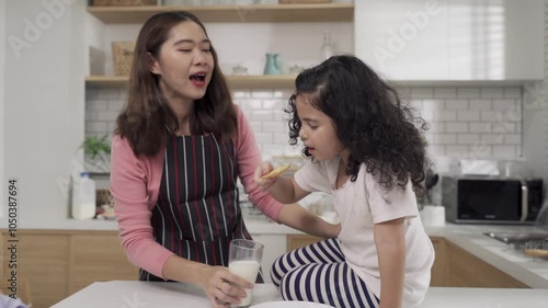 A woman is looking at a little girl is drinking milk and eating cookies deliciously. The woman is wearing an apron and the girl is sitting on a chair.