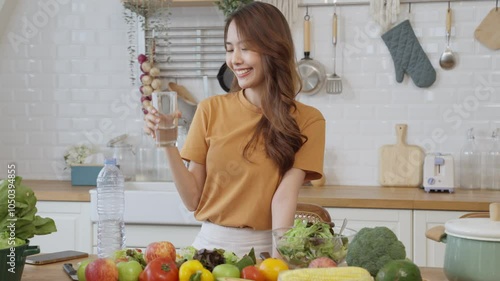 A woman is drinking water while standing in front of a table full of fruits and vegetables. The table is covered with a variety of fruits and vegetables, including apples, oranges, and broccoli