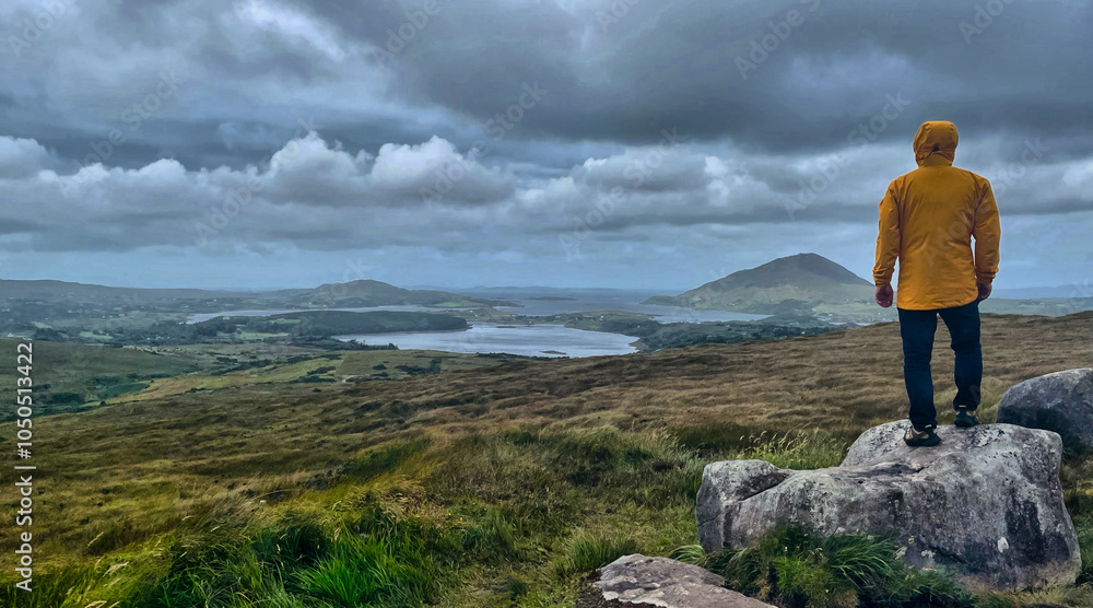 Wanderer (männlich) mit gelber Regenjacke von hinten auf einem Fels mit Panorama Blick zur Küste am Lower Diamond Hill im Connemara National Park, Irland Wanderer (männlich) mit gelber Regenjacke von hinten auf einem Fels mit Panorama Blick zur Küste am Lower Diamond Hill im Connemara National Park, Irland