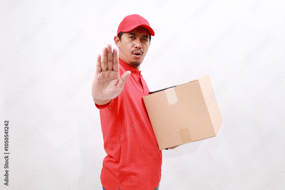 Asian delivery courier in red t-shirt and cap carrying a box package while making stop gesture showing palm of hand. isolated over white background Asian delivery courier in red t-shirt and cap carrying a box package while making stop gesture showing palm of hand. isolated over white background