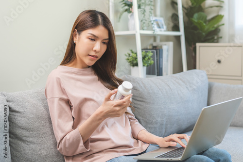 Wellness and dieting asian young woman, girl working from home using computer, typing or searching prescription on medicine label about vitamins information online, holding bottle of food supplement.