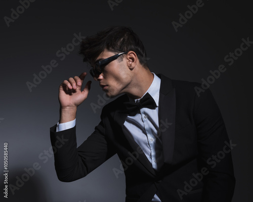 elegant young groom in black tuxedo looking to side and fixing sunglasses