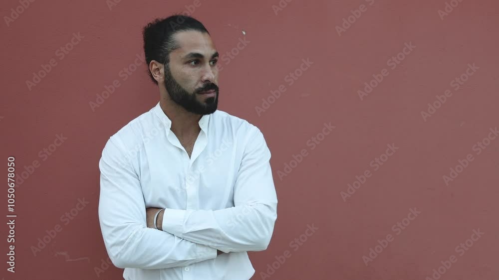 A bearded dark-skinned handsome guy in a white shirt poses against a red wall in the evening. Camera movement from left to right. A bearded dark-skinned handsome guy in a white shirt poses against a red wall in the evening. Camera movement from left to right.
