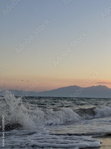 Waves crashing gently on the shore at sunset, with mountains in the background near the coast