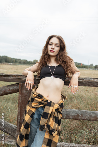 Beautiful fashionable rural girl in fashionable clothes with shirt, top and jeans stands near wooden fence countryside