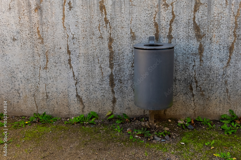 Obraz premium A gray trash can stands against a textured wall, with green plants growing at its base