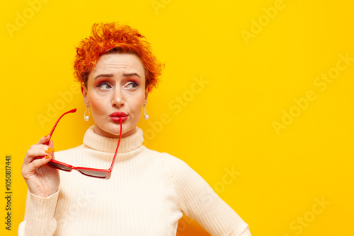 young red-haired uncertain woman with curly hair holds sunglasses and thinks on a yellow isolated background, puzzled girl with beautiful make-up doubts and plans and imagines