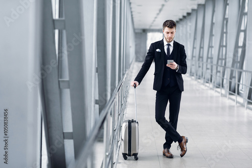 Portrait of confident guy in suit using cell phone standing in airport lobby holding trolley bag, copy space, full length