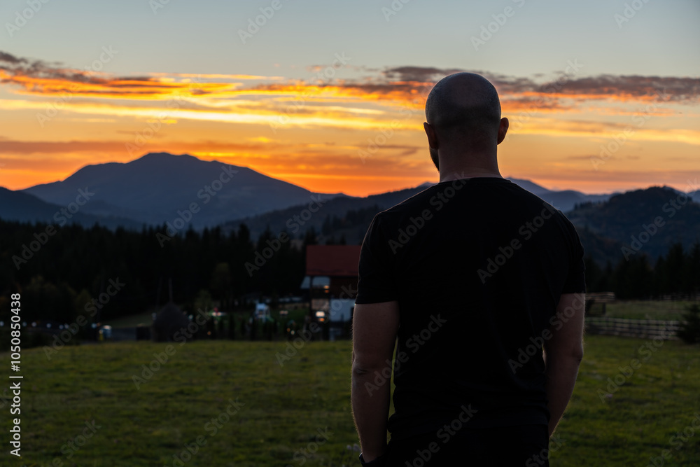 Fototapeta premium A man stands with his back to the camera, gazing at the sunset over mountains in a peaceful rural setting, creating a contemplative mood.