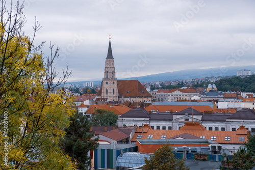 Scenic view of a historic church and rooftops in Cluj-Napoca, Romania, surrounded by autumn trees and a cloudy sky.