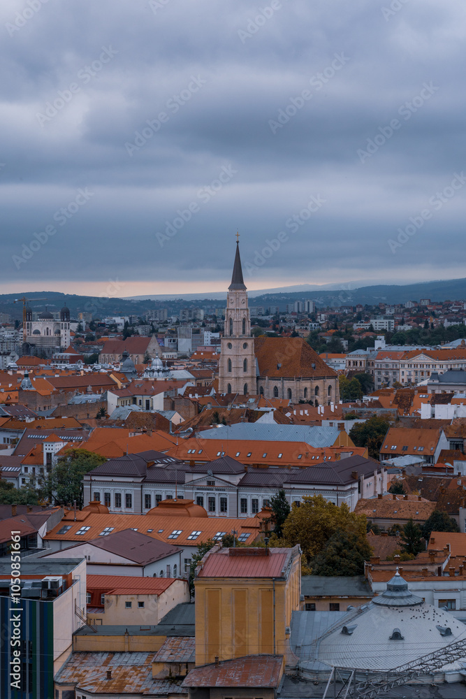 Obraz premium Panoramic view of Cluj-Napoca featuring St. Michael's Church and surrounding rooftops under a cloudy evening sky.