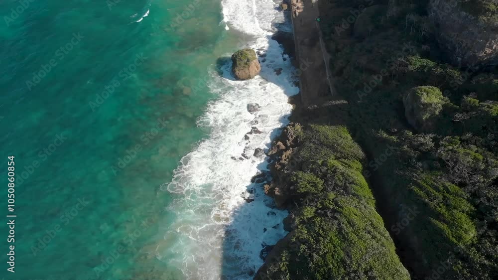 An aerial view shows a rocky coastline with turquoise ocean waves crashing against the shore, surrounded by lush green vegetation in Isabela, Tunel of Guajataca, Puerto Rico. An aerial view shows a rocky coastline with turquoise ocean waves crashing against the shore, surrounded by lush green vegetation in Isabela, Tunel of Guajataca, Puerto Rico.