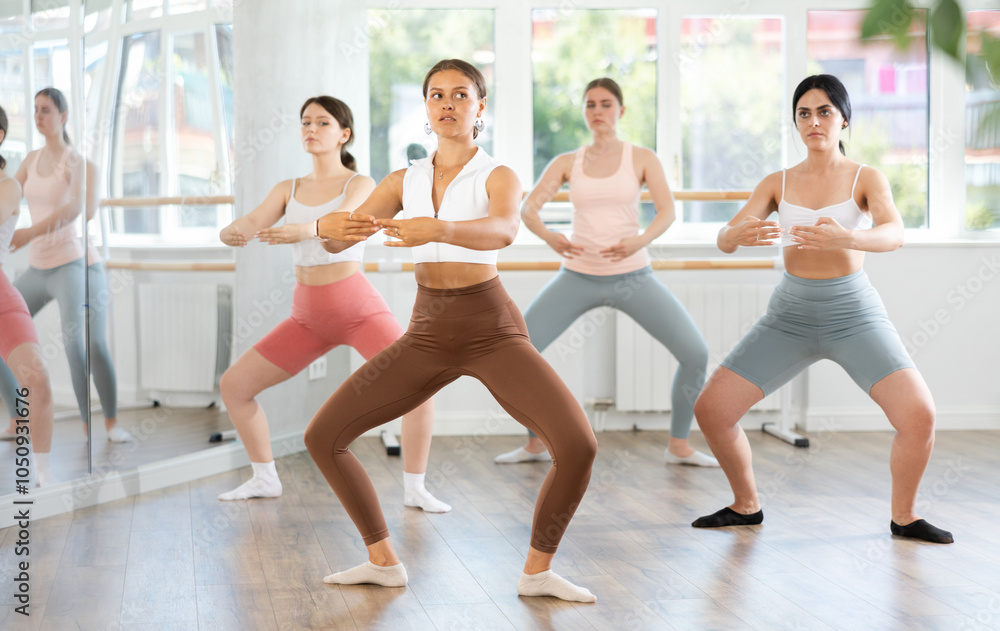Obraz premium Graceful young woman in sportswear confidently leading ballet class with female amateur dancers following her movements in spacious serene dance studio
