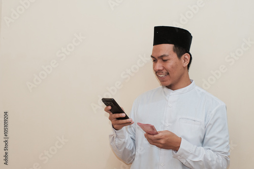 Smiling Southeast Asian Muslim man holding Rupiah banknotes while looking at his mobile phone. Ideal for finance and lifestyle concepts. People religious Islam lifestyle concept. celebrating Ramadan.