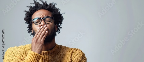 A young man wearing a yellow sweater yawns with a tired expression, conveying feelings of sleepiness and relaxation in a modern indoor setting.