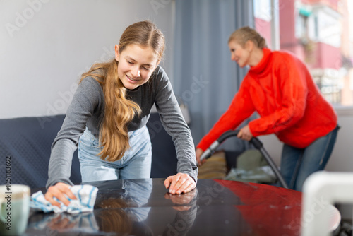 Hardworking daughter cleans the table with a rag while mother vacuums the room photo