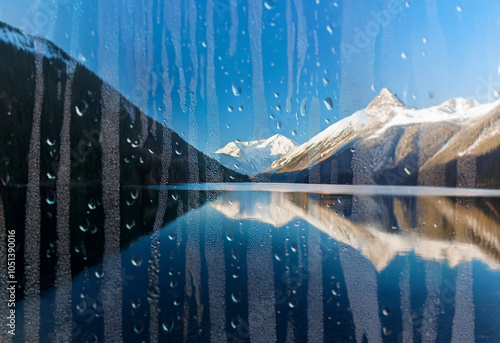 A view of snow-capped mountains reflected in a still lake seen through a rain-streaked window.