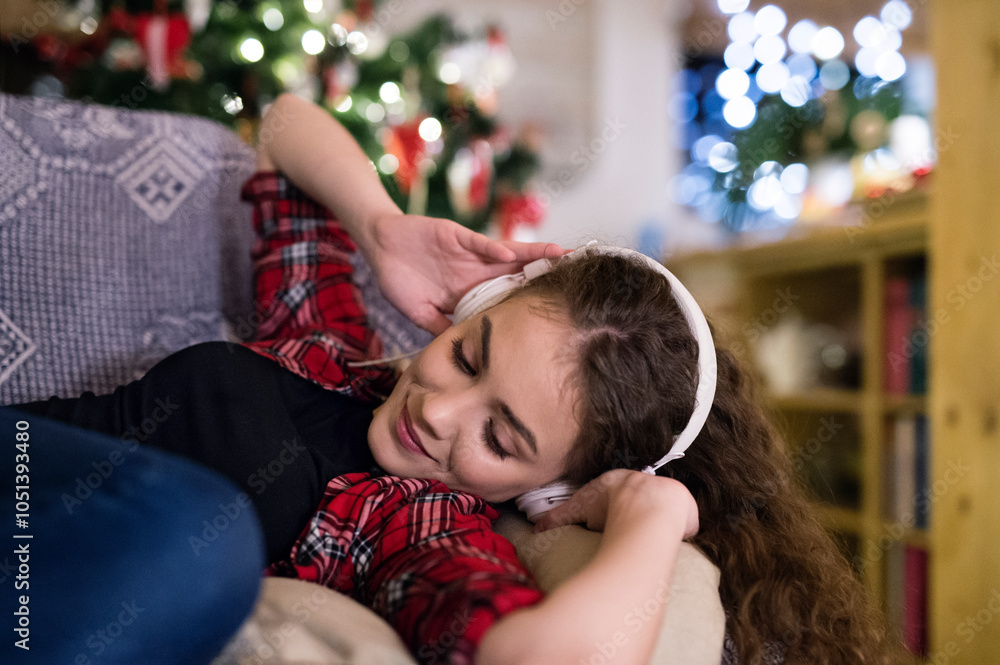 Fototapeta premium Student having hygge Christmas at home. Woman is sitting on sofa by Christmas tree, listening music with headphones.