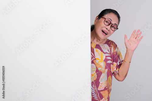 Elderly Asian woman peeking and waving her hand from behind a wall with happy expression photo