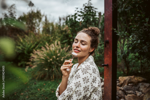 Beautiful woman massaging her face with rose quartz roller, sitting in garden. Outdoor skin care routine for young woman. photo