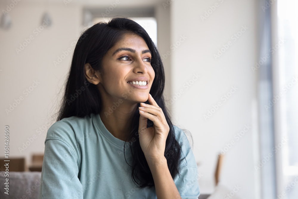 Happy daydreaming young Indian woman posing at home, looking away with toothy smile, touching chin, facial skin, sitting on couch, thinking, dreaming, enjoying homey comfort Happy daydreaming young Indian woman posing at home, looking away with toothy smile, touching chin, facial skin, sitting on couch, thinking, dreaming, enjoying homey comfort