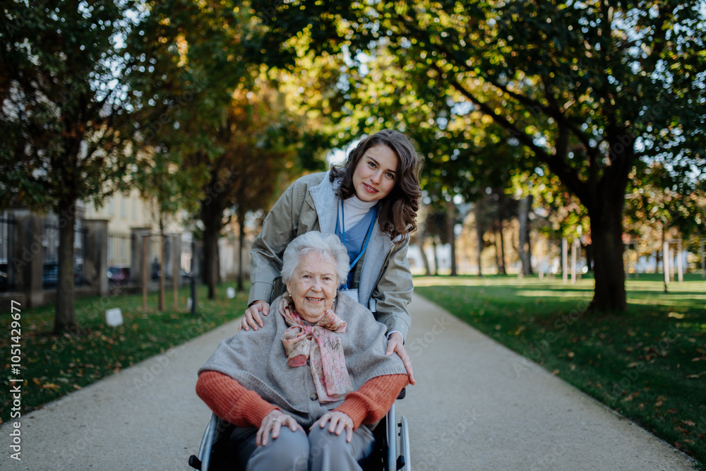 Fototapeta premium Disabled elderly woman in wheelchair on walk in the park during warm autumn day. Young nurse pushing wheelchair, talking with senior woman.