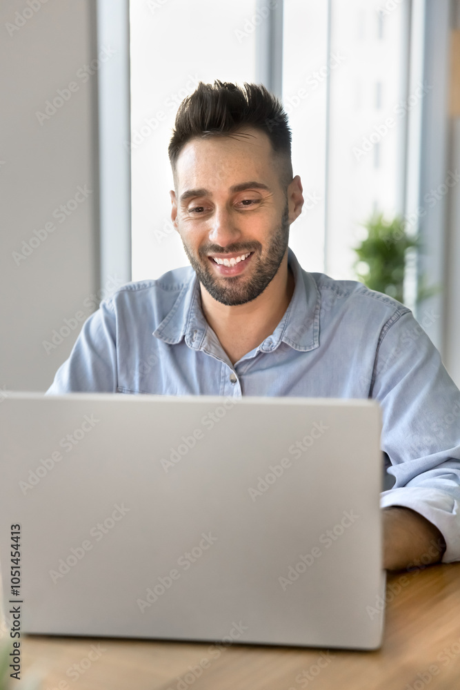 Fototapeta premium Positive handsome freelance professional man in casual working on online project at laptop, typing, smiling, browsing Internet, social media, sitting at workplace table in home office. Vertical shot