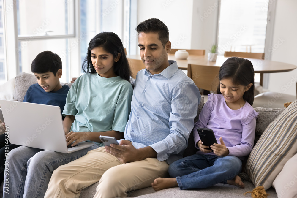 Couple of internet addicted Indian parents and two little sibling kids focused on gadgets ignoring each other, sitting close on home couch, using smartphones, laptop, playing online game Couple of internet addicted Indian parents and two little sibling kids focused on gadgets ignoring each other, sitting close on home couch, using smartphones, laptop, playing online game