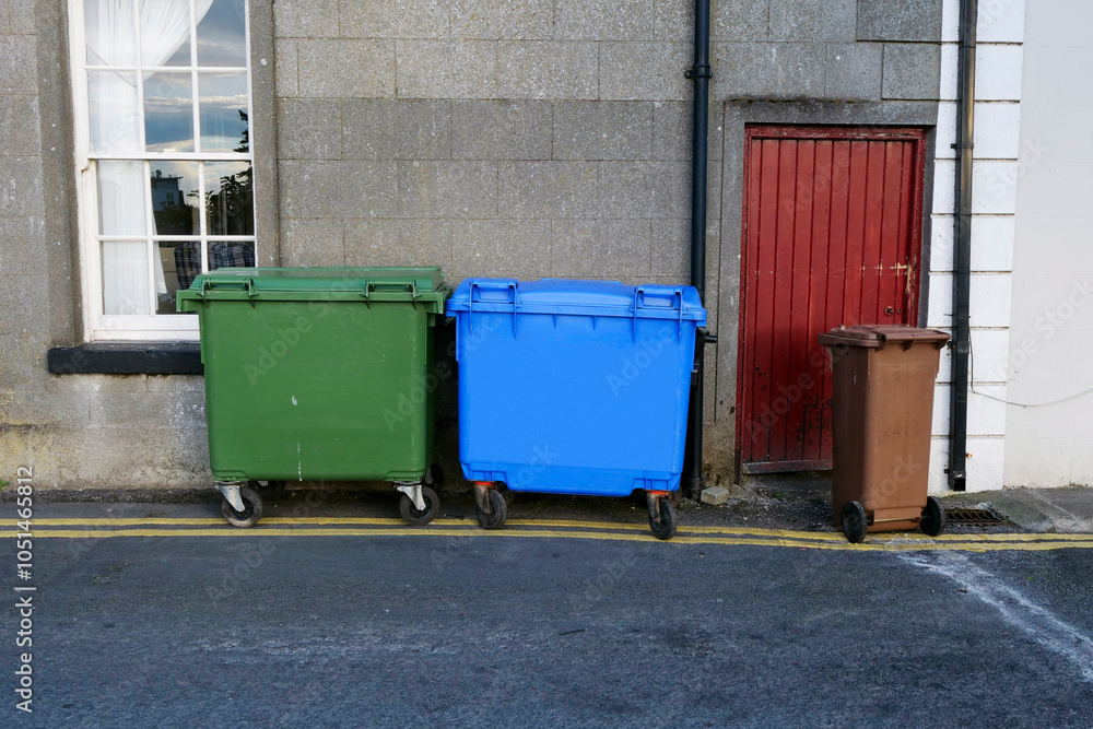 Three trash cans are lined up on the sidewalk. One is green, one is blue, and one is brown. Waste and ecology management in a city. Household job. Excess rubbish with modern consumer habit behavior. Three trash cans are lined up on the sidewalk. One is green, one is blue, and one is brown. Waste and ecology management in a city. Household job. Excess rubbish with modern consumer habit behavior.