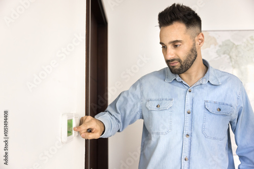 Handsome young Hispanic homeowner man touching wall mounted panel, smart home gadget, activating air conditioner, thermostat, alarm system, regulating air temperature in apartment