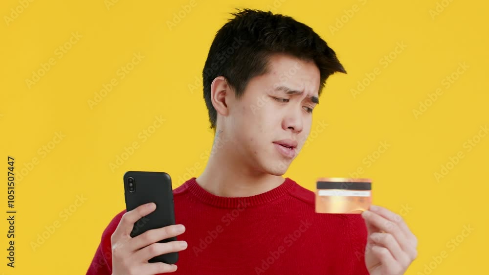 Asian young man in a red sweater holds a credit card in one hand and a smartphone in the other, displaying a thoughtful expression while engaged in an online transaction. Asian young man in a red sweater holds a credit card in one hand and a smartphone in the other, displaying a thoughtful expression while engaged in an online transaction.