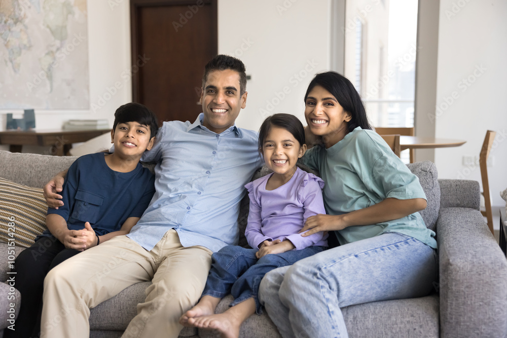Happy Indian parents and two little preteen children sitting together on home couch, looking at camera, smiling, laughing, enjoying homey comfort, closeness, posing for family portrait Happy Indian parents and two little preteen children sitting together on home couch, looking at camera, smiling, laughing, enjoying homey comfort, closeness, posing for family portrait