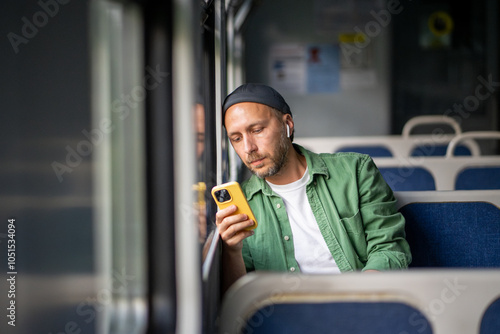 Thoughtful man concentrating on mobile phone rides on electric train reading e-book, scrolling social media, listens to music on headphones to pass time, leaning to window. Public transport trip alone