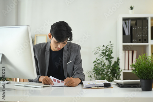 A man in a suit is sitting at a desk with a computer monitor and a notebook