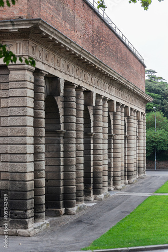 Wallpaper Mural A grand colonnade with robust, textured stone pillars stands in quiet strength. The repetitive arches create a rhythmic architectural pattern against a high brick wall, surrounded by greenery. Torontodigital.ca