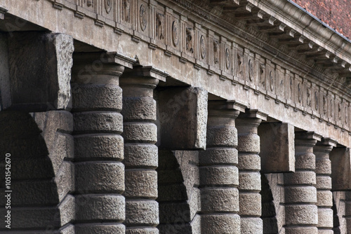 Wallpaper Mural A grand colonnade with robust, textured stone pillars stands in quiet strength. The repetitive arches create a rhythmic architectural pattern against a high brick wall, surrounded by greenery. Torontodigital.ca