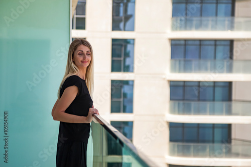 Portrait of perfect blonde woman in black attire on balcony of skyscraper Dubai UAE, smile looking away. Lovely lady posing on terrace of tower block. Luxury chic lifestyle concept. Copy ad text space