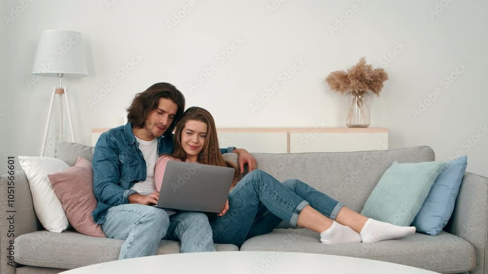 A young couple sits together on a comfortable couch, sharing a laptop. They are relaxed, surrounded by soft cushions, and seem engaged in conversation and laughter. A young couple sits together on a comfortable couch, sharing a laptop. They are relaxed, surrounded by soft cushions, and seem engaged in conversation and laughter.