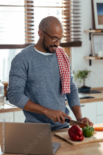 Smiling man cutting vegetable and learning recipe through laptop at home photo