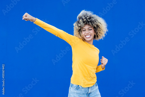 Cheerful woman with curly hair holding fist against blue background photo