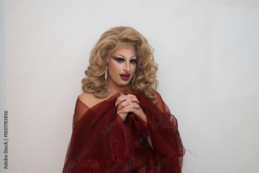 Fototapeta premium A drag queen with blonde wig and luxurious earrings and covered in red silk and on a white background with clasped hands praying imitating the virgin. Concept of lgtbqi rights and equality.